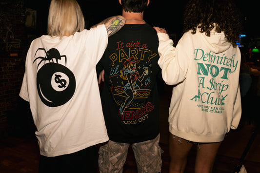 Three people standing together with their backs to the camera, wearing oversized graphic t-shirts and a hoodie with retro-style back prints. The left person wears a white oversized t-shirt with a black pool billiards and spider graphic.