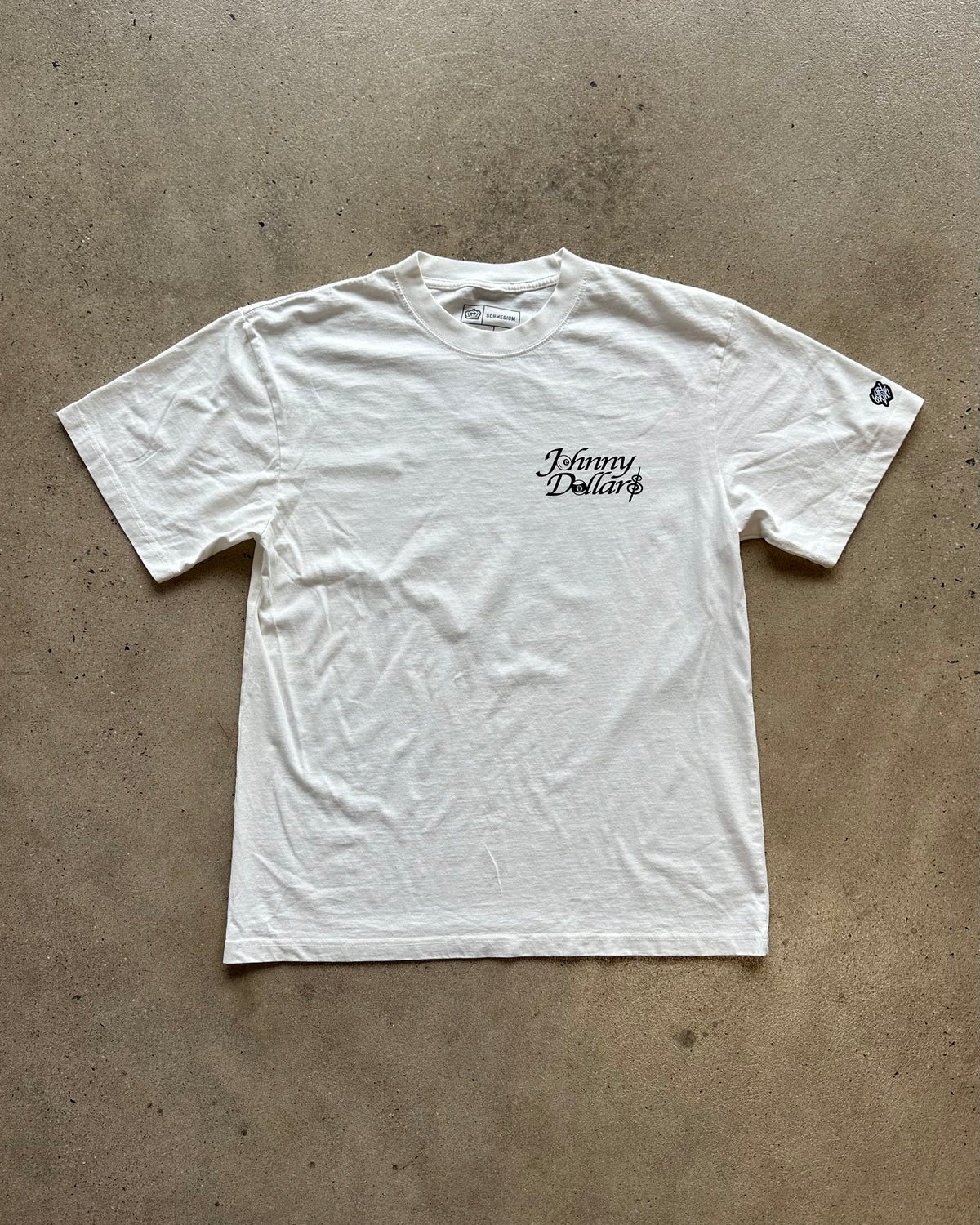 A white short-sleeve t-shirt with 'Johnny Dollar' printed in black script on the left chest, laid flat on a textured concrete floor.