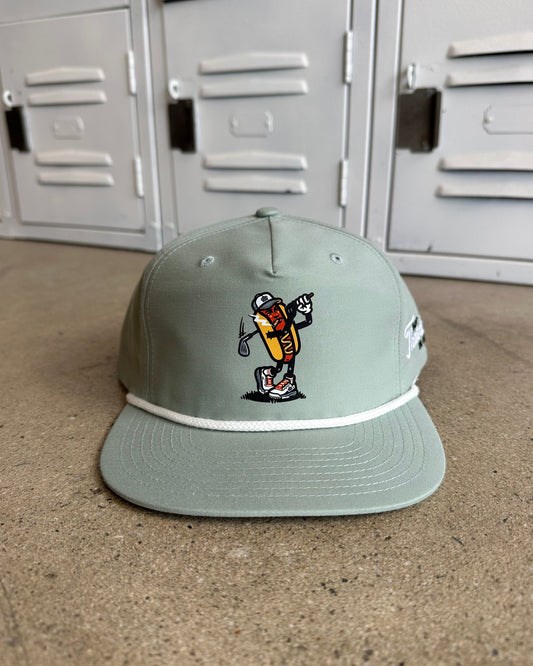A sage green 5-panel cap with a white rope detail and a cartoon mascot embroidered on the front, resting on a concrete floor in front of gray metal lockers.