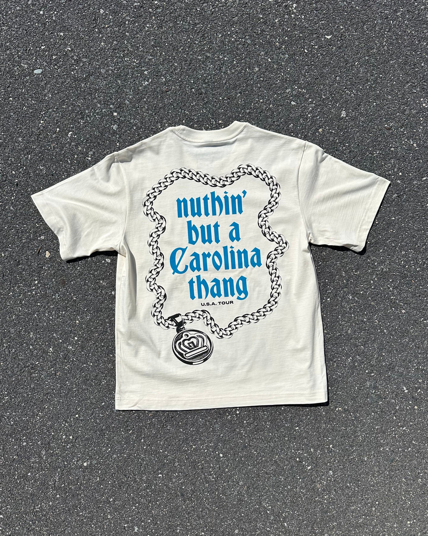 A white graphic t-shirt laid flat on dark asphalt pavement. The back of the shirt features a blue and black graphic of a rose and chain with the text 'nuthin' but a Carolina thang' in blue.