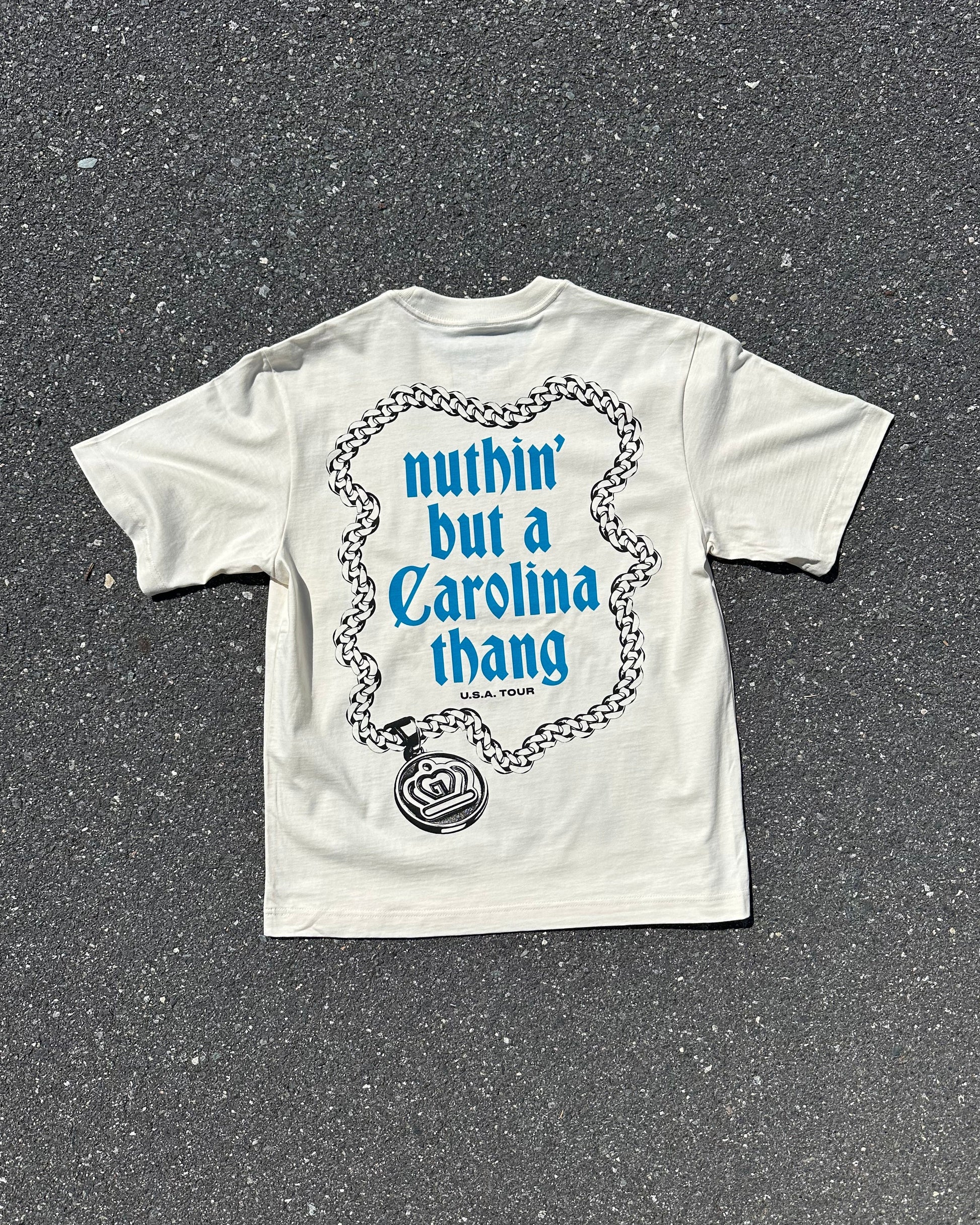 A white graphic t-shirt laid flat on dark asphalt pavement. The back of the shirt features a blue and black graphic of a rose and chain with the text 'nuthin' but a Carolina thang' in blue.