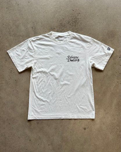A white short-sleeve t-shirt with 'Johnny Dollar' printed in black script on the left chest, laid flat on a textured concrete floor.