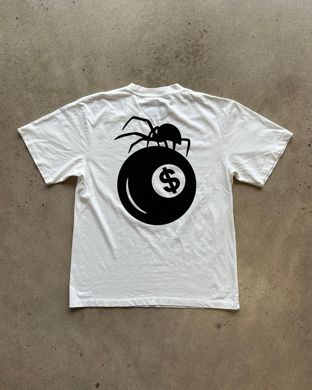 A white short-sleeve t-shirt laid flat on a textured concrete floor, featuring a large black graphic on the back of a spider perched on a billiards ball with a dollar sign.
