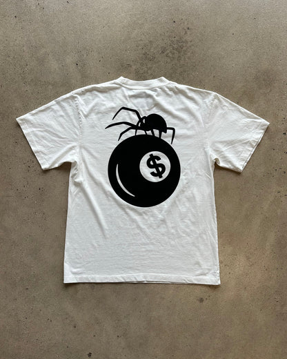 A white short-sleeve t-shirt laid flat on a textured concrete floor, featuring a large black graphic on the back of a spider perched on a billiards ball with a dollar sign.