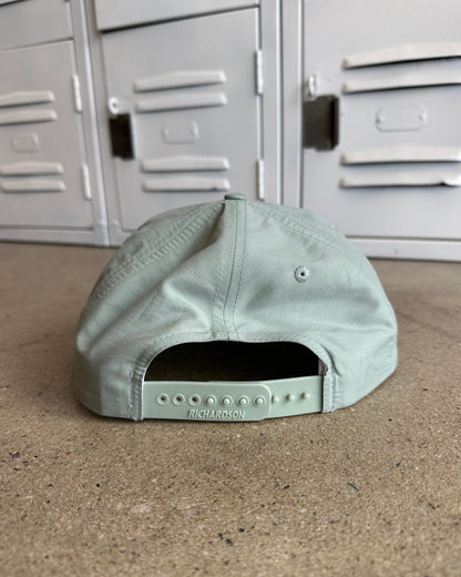 A sage green baseball cap viewed from the back, resting on a concrete floor in front of gray metal lockers. The brand name 'RICHARDSON' is debossed on the back strap.