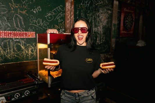 A woman wearing red-tinted sunglasses and a black graphic t-shirt stands in a dimly lit restaurant or bar setting, holding two hot dogs in her hands. The background features chalkboard walls with drawings and a hot dog grill.