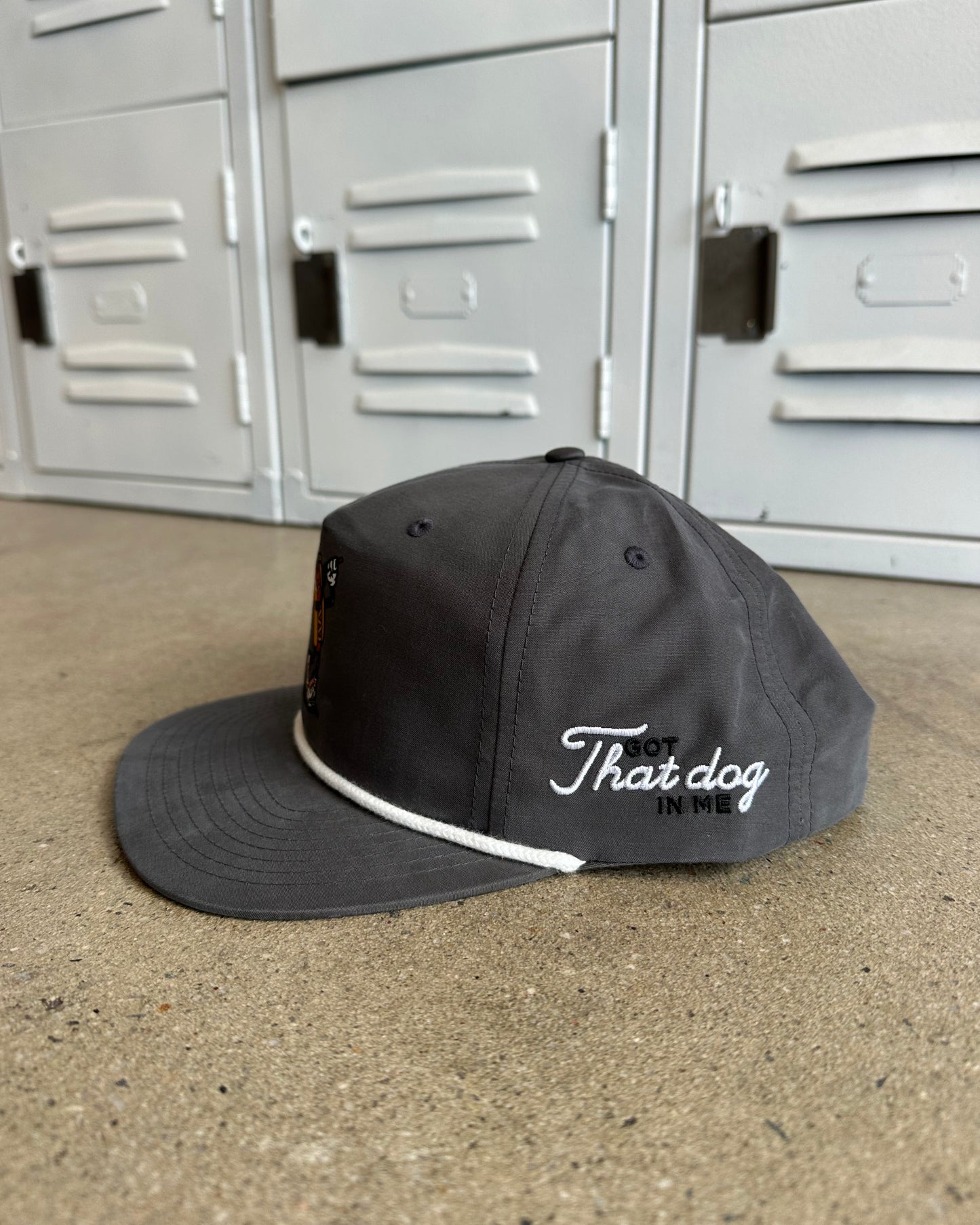 A dark gray flat-brim cap with white embroidered text reading 'That dog' and a white piping detail along the edge of the brim, resting on a concrete floor in front of gray metal lockers.