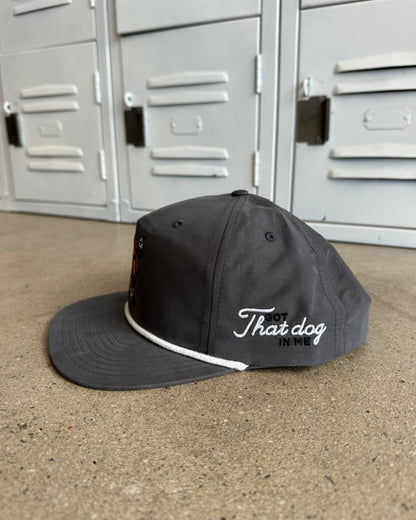 A dark gray flat-brim cap with white embroidered text reading 'That dog' and a white piping detail along the edge of the brim, resting on a concrete floor in front of gray metal lockers.