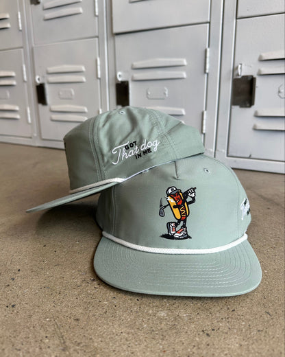 A sage green snapback cap with embroidered details, featuring a hot dog character on the front and 'The Dog' script on the side, resting on a concrete floor in front of gray metal lockers.
