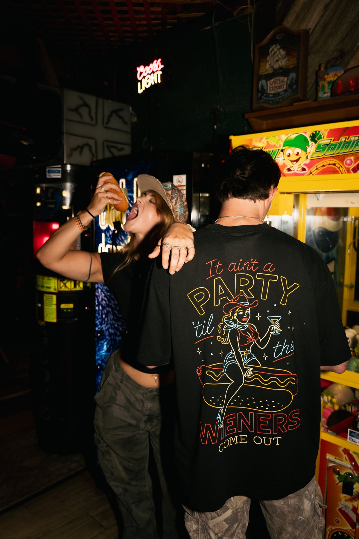 A person wearing a black graphic t-shirt with a large retro-style party graphic on the back, standing in a dimly lit bar or arcade environment next to another person.
