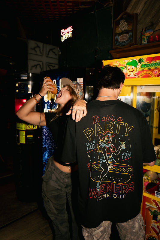 A person wearing a black graphic t-shirt with a large retro-style party graphic on the back, standing in a dimly lit bar or arcade environment next to another person.