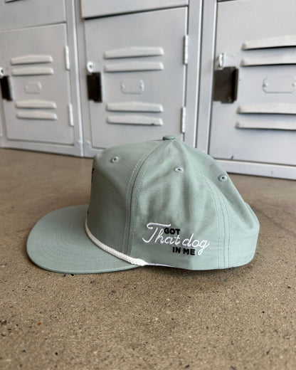 A sage green flat-brim cap with white embroidered text reading 'That dog in my' resting on a concrete floor in front of gray metal lockers.
