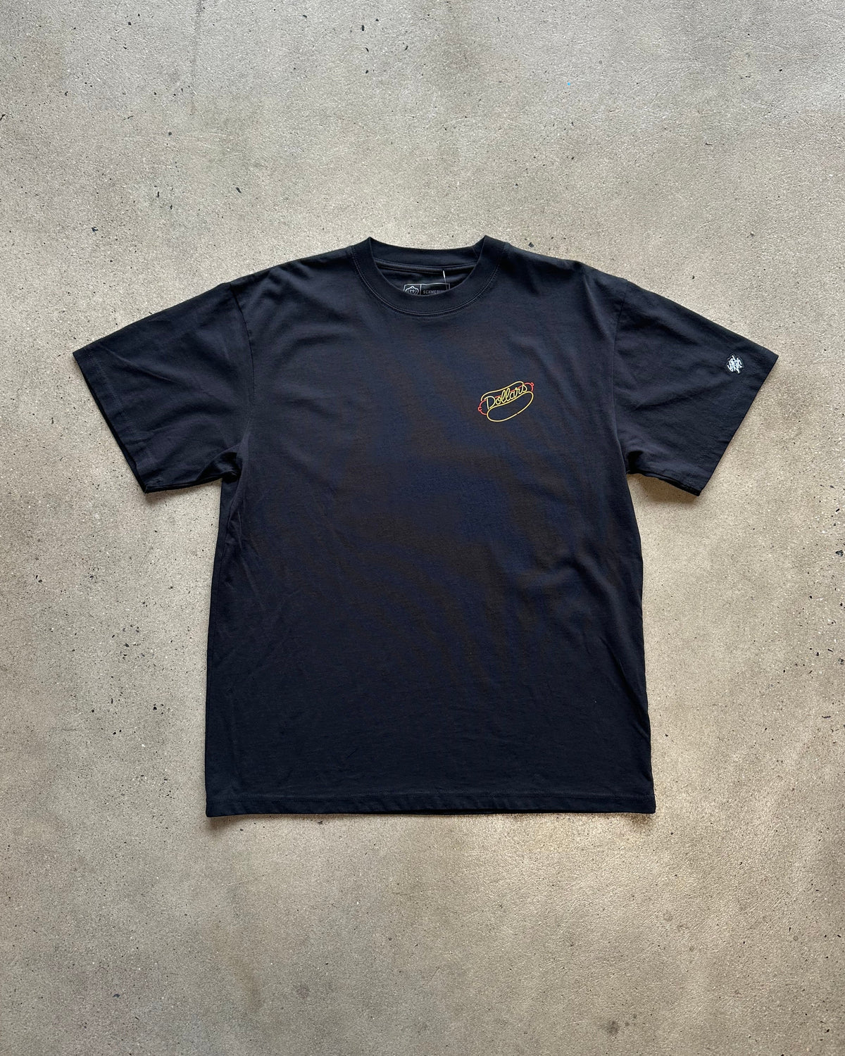 A black short-sleeve t-shirt with a small yellow and white graphic on the left chest, laid flat on a concrete floor. A white sneaker is partially visible at the bottom edge.