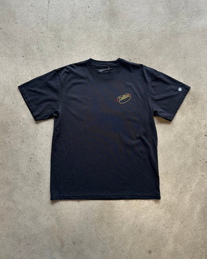 A black short-sleeve t-shirt with a small yellow and white graphic on the left chest, laid flat on a concrete floor. A white sneaker is partially visible at the bottom edge.
