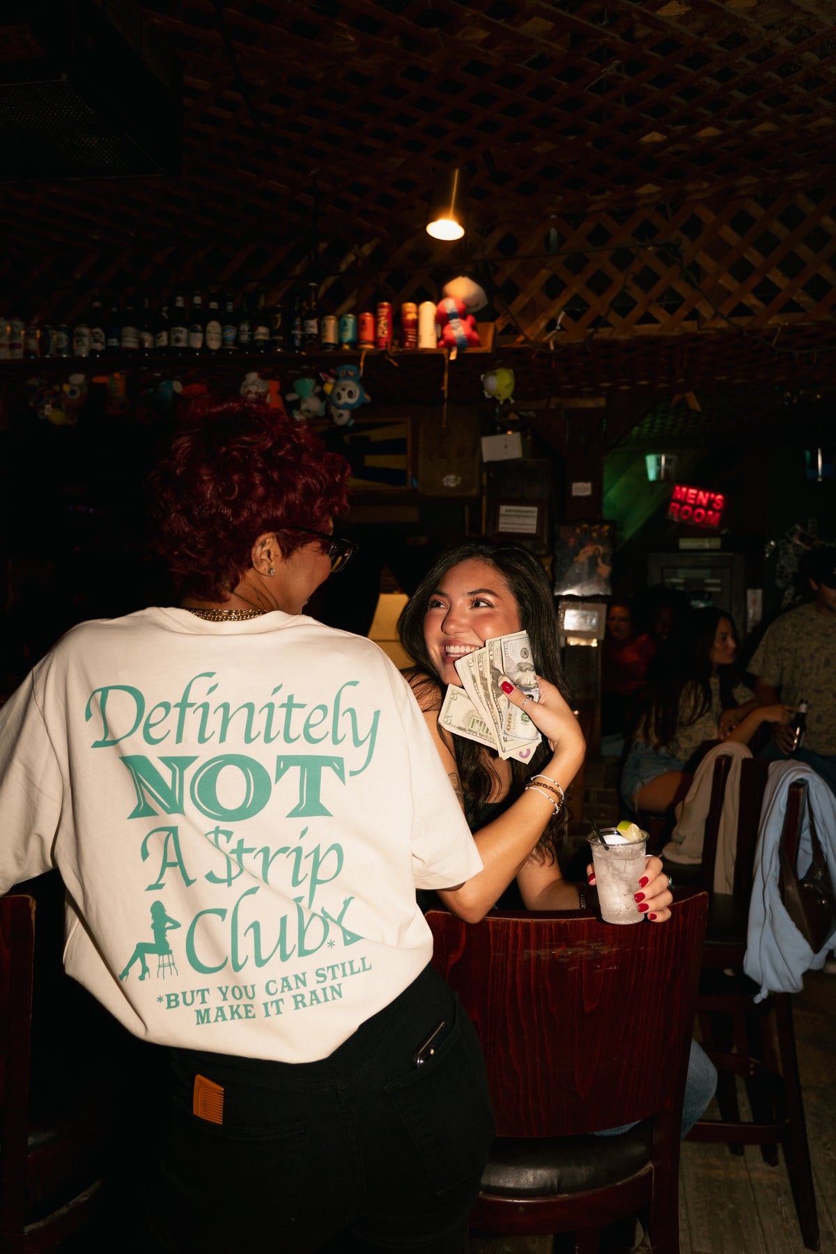 A person standing in a dimly lit bar or club, shown from the back wearing a white graphic T-shirt with green text that reads 'Definitely NOT A Strip Chub' and 'BUT YOU CAN STILL MAKE IT RAIN.' Another person seated at the table is visible in the background holding cash.