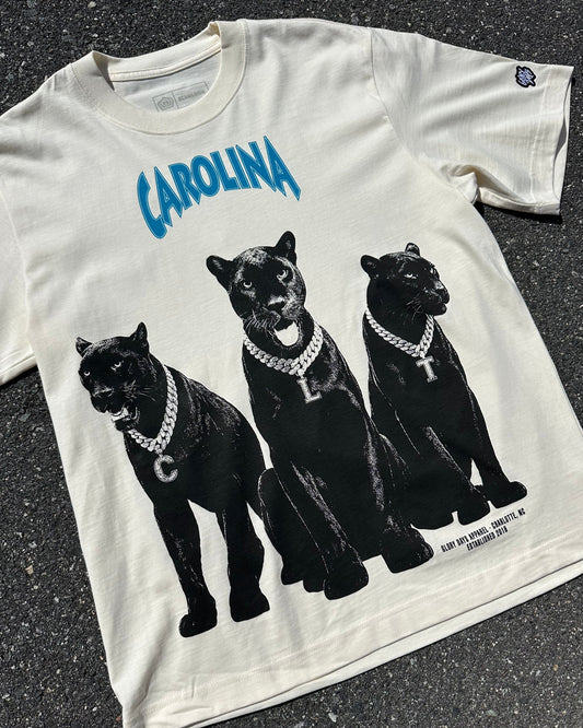 An off-white short-sleeve graphic t-shirt laid flat on asphalt. The shirt features a large black graphic of three panthers wearing gold chains, with the word 'CAROLINA' printed in blue above them.
