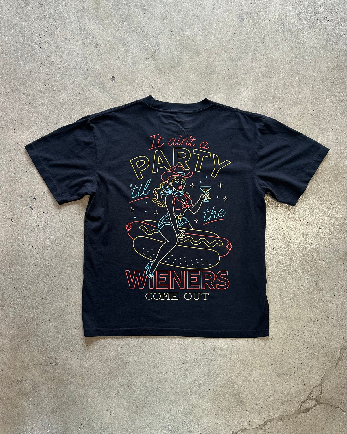 A navy blue graphic t-shirt laid flat on a textured concrete surface. The shirt features a retro-style illustration of a pin-up girl sitting on a hot dog with the text 'It ain't a PARTY til the WIENERS COME OUT' printed in yellow, red, and light blue.