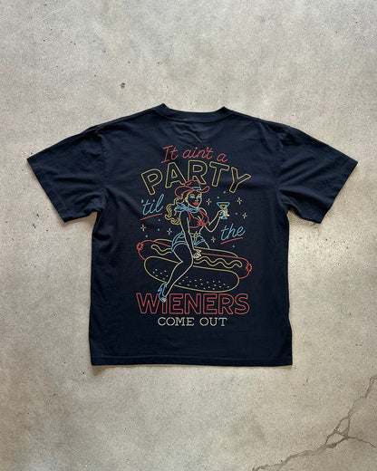 A navy blue graphic t-shirt laid flat on a textured concrete surface. The shirt features a retro-style illustration of a pin-up girl sitting on a hot dog with the text 'It ain't a PARTY til the WIENERS COME OUT' printed in yellow, red, and light blue.