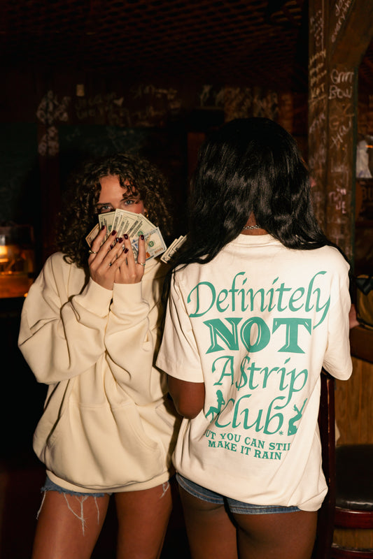 Two women standing in a dimly lit bar or club. The woman on the left wears an off-white hoodie and holds cash near her face.