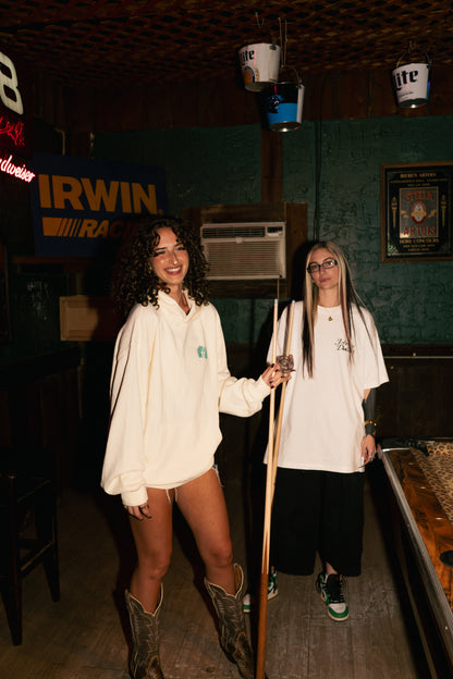 Two women standing in a dimly lit bar or lounge setting, both wearing oversized white graphic t-shirts. The woman on the left is wearing brown cowboy boots, and the woman on the right is wearing black pants.