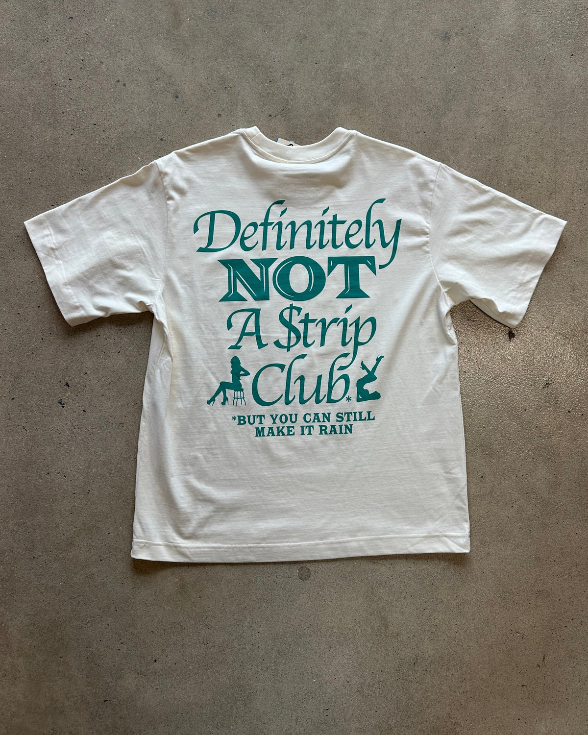 A white graphic t-shirt laid flat on a textured concrete floor, featuring a teal green text print that reads 'Definitely NOT A Strip Club' with a small dancer silhouette and the quote 'BUT YOU CAN STILL MAKE IT RAIN' underneath. A second plain white t-shirt is partially visible at the top edge of the frame.