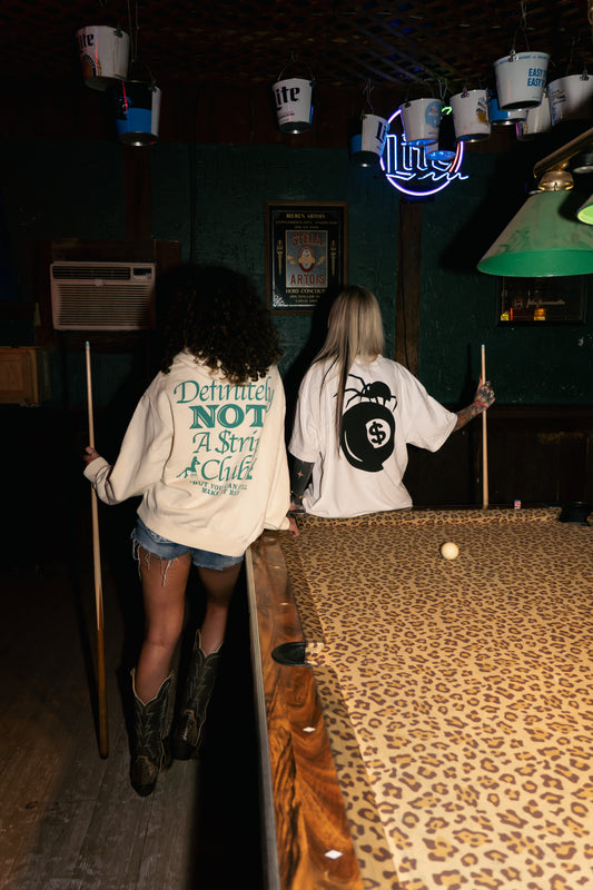 Two women standing in a dimly lit bar or pool room, seen from behind, wearing oversized white graphic sweatshirts. The sweatshirt on the left has teal text reading 'Definitely Not A Star A Club,' and the sweatshirt on the right has a large black circular graphic.