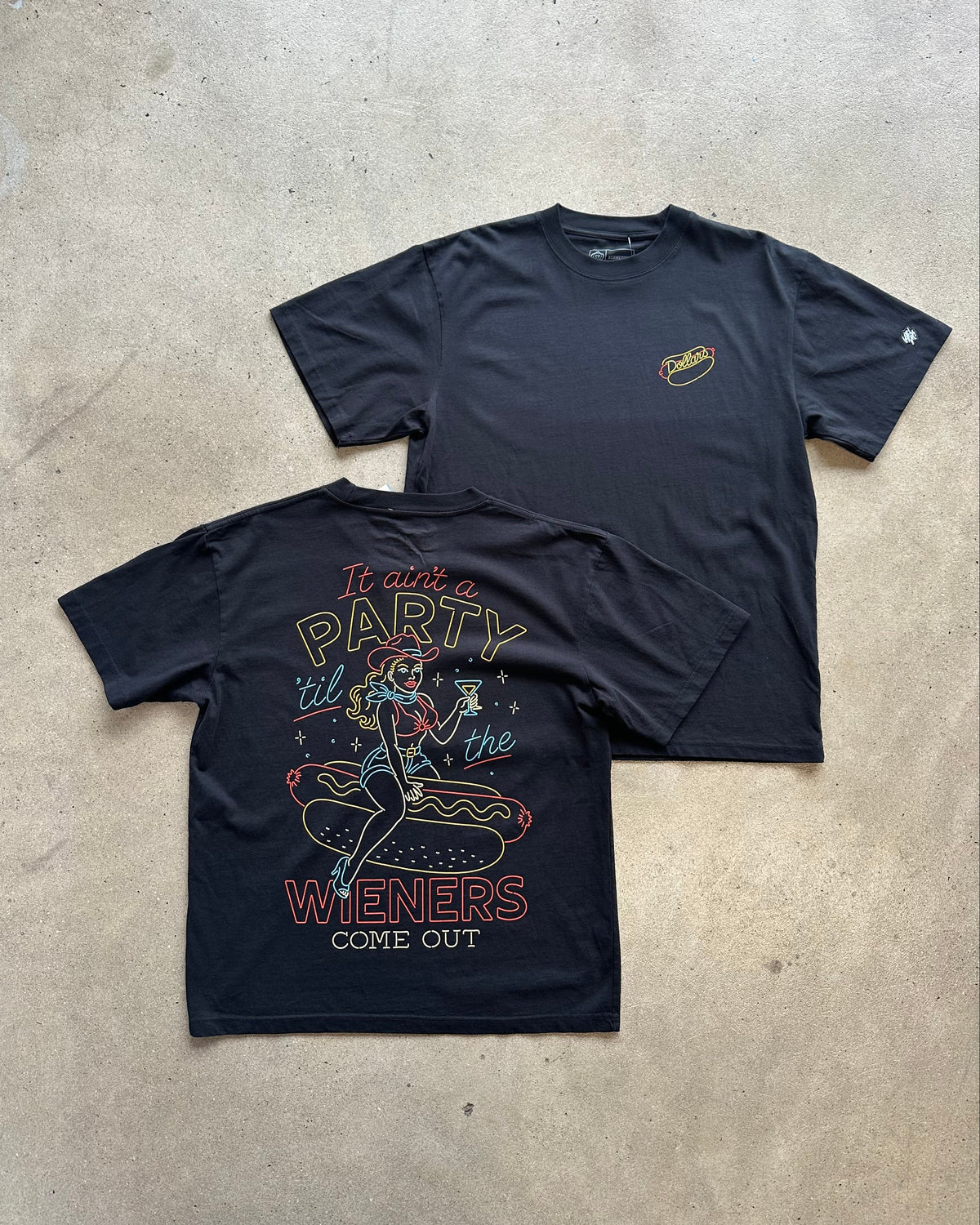 Two black graphic t-shirts laid flat on a concrete floor. The front shirt shows a small yellow graphic on the left chest, and the back shirt features a large colorful retro graphic with the text 'It ain't a PARTY it's the WIENERS COME OUT' and an illustration of a woman eating a hot dog.