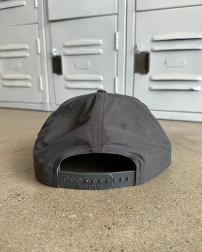 A dark gray baseball cap viewed from the back, resting on a concrete floor in front of gray metal lockers. The brand name 'RICHARDSON' is visible on the back strap.