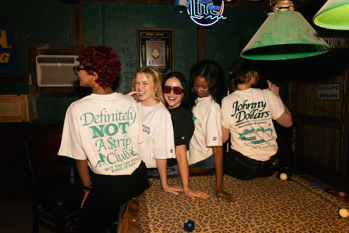 Four women standing in a dimly lit bar or club interior, all wearing matching off-white graphic T-shirts with large teal text on the back. The shirts read phrases including 'Definitely NOT A Strip Club' and 'Johnny Dollars'.