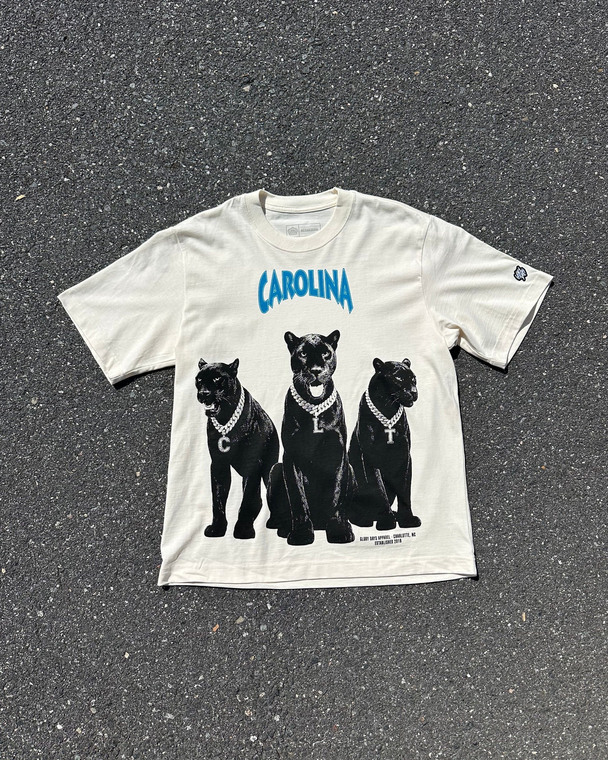 An off-white graphic t-shirt laid flat on dark asphalt. The shirt features the word 'CAROLINA' in blue arched text above a graphic of three black panthers wearing silver chains.