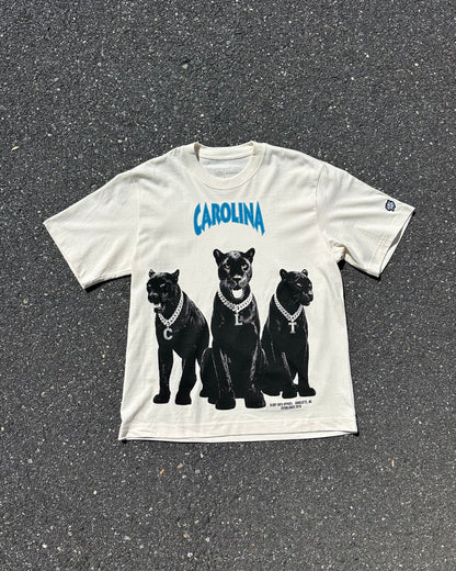 An off-white graphic t-shirt laid flat on dark asphalt. The shirt features the word 'CAROLINA' in blue arched text above a graphic of three black panthers wearing silver chains.