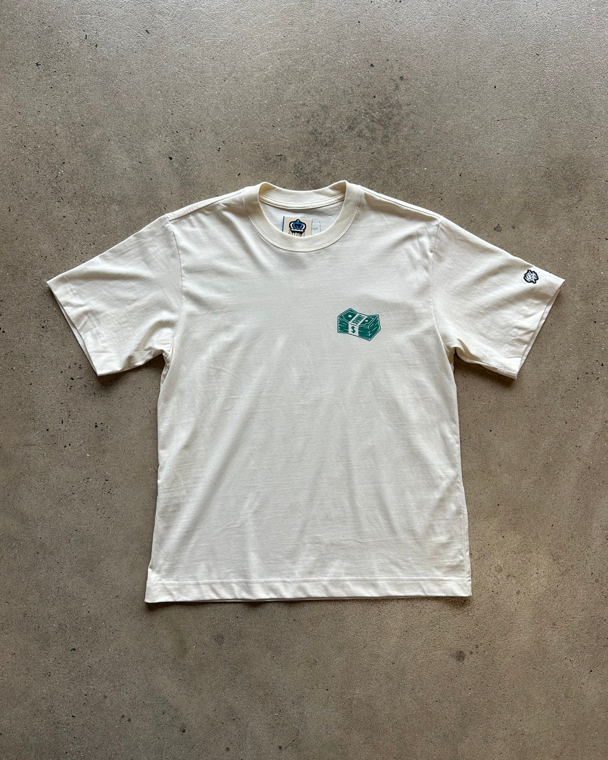 An off-white short-sleeve t-shirt laid flat on a concrete floor, featuring a small green graphic on the left chest and a small circular logo on the right sleeve.
