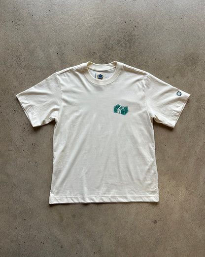 An off-white short-sleeve t-shirt laid flat on a concrete floor, featuring a small green graphic on the left chest and a small circular logo on the right sleeve.