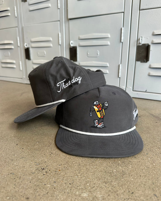 Two dark gray flat-brim capns stacked on a concrete floor in front of gray metal lockers. The front cap features a colorful embroidered hot dog character and a white piping accent, while the back cap displays the white cursive text 'That dog'.