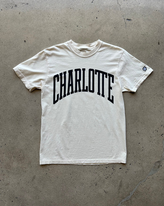 White t-shirt with 'Charlotte' printed on it, lying flat on a concrete surface.